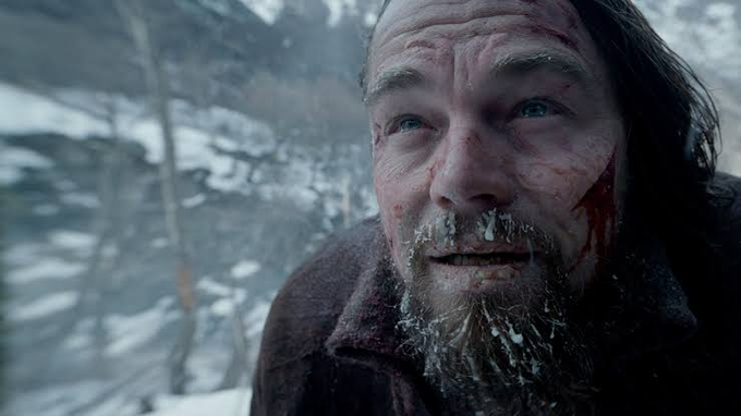 A close-up of Leonardo DiCaprio with a bloodied face and frost-covered beard, smiling slightly in a snowy setting.