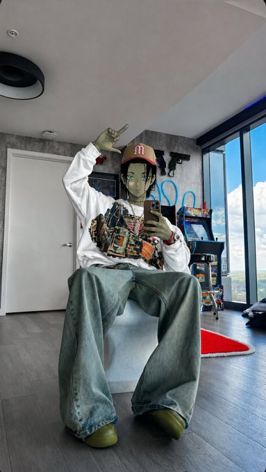 A person sitting on a chair taking a mirror selfie with their phone, wearing a McDonald's-themed hat, a white sweatshirt with graphics, baggy jeans, and green shoes, with arcade machines visible in the background.