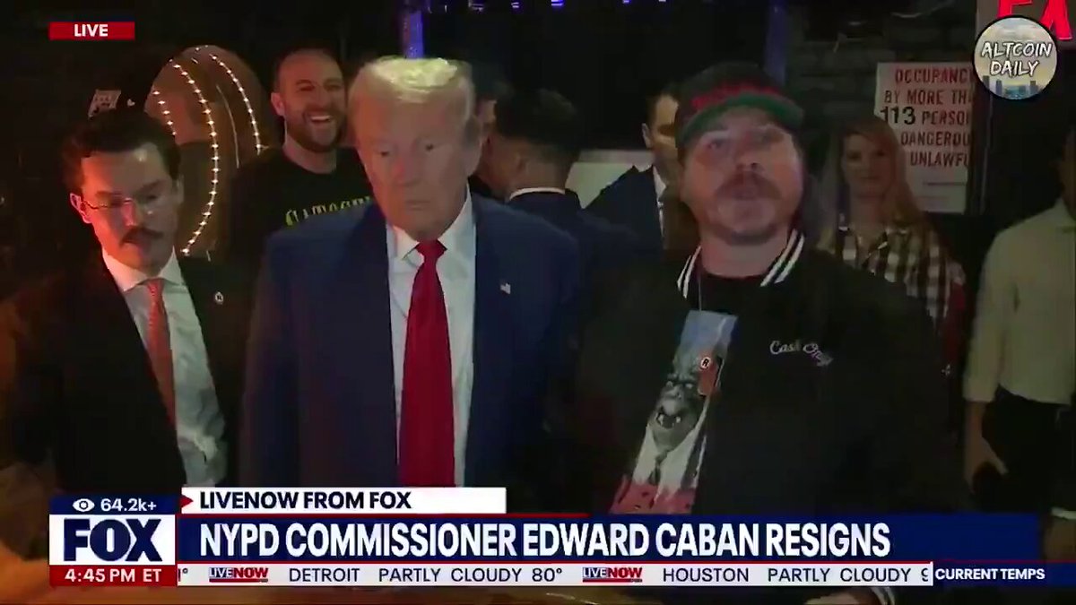 The image shows a group of people, including two men wearing ties, standing around each other at an event. One man is wearing a hat that has Donald Trump's face on it. The other man is wearing a shirt with a picture of Edward Caban on it. They are surrounded by several other people who appear to be enjoying the gathering as well. In the background, there is a TV screen displaying news about Bitcoin transactions involving President Trump and another person.