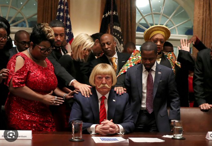A staged comedic photograph depicting a Trump lookalike seated at a conference table with hands resting on it, while several people stand behind him with their hands placed on his shoulders in a prayer-like gesture, with American flags in the background.