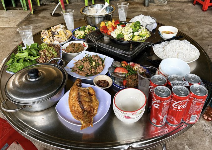 A round metal table is laden with a feast of Asian cuisine, featuring a whole fried fish, grilled meat, vegetables, rice noodles, and several cans of Coca-Cola.