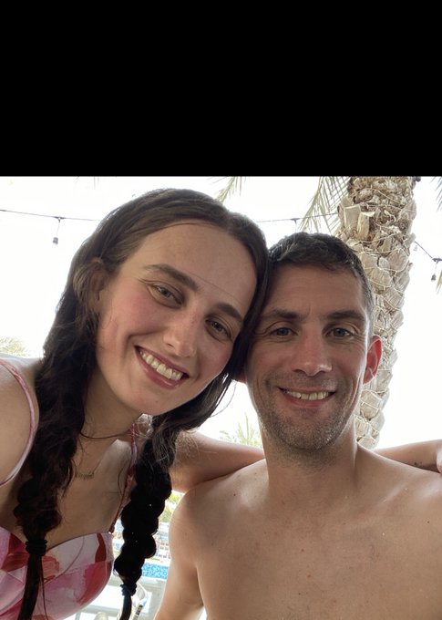 A selfie of a smiling couple—a woman with long dark braided hair and a shirtless man—posed closely together in an outdoor setting with palm trees in the background.