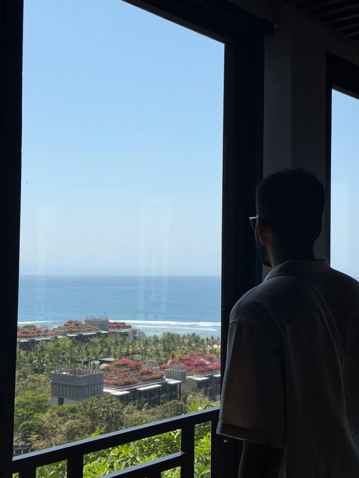A silhouetted man wearing glasses and a white shirt stands looking out a large window at an ocean view with coastline, buildings, and flowering trees below.