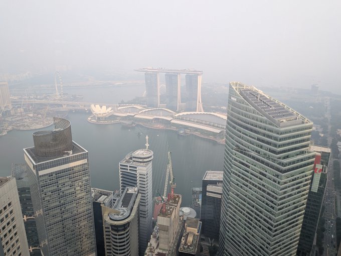 The image shows Singapore's Marina Bay skyline shrouded in haze, with the iconic Marina Bay Sands hotel complex visible across the water from a high-rise vantage point. This smoky, atmospheric view captures the city as it recovers from a period of haze, likely caused by regional forest fires, adding a moody tone to the post about "recovering from all the greatness."