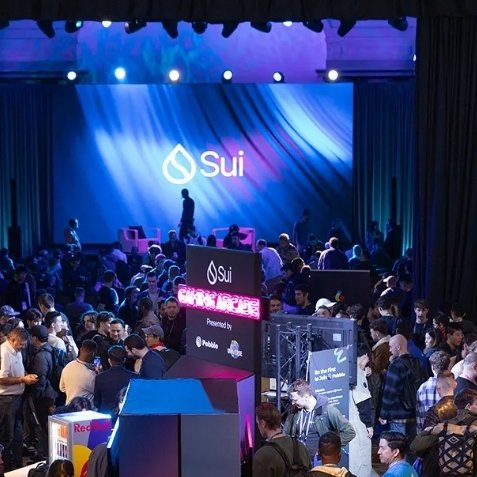A crowded Sui cryptocurrency conference event with the Sui logo prominently displayed on a large blue screen backdrop and on signage within the venue.