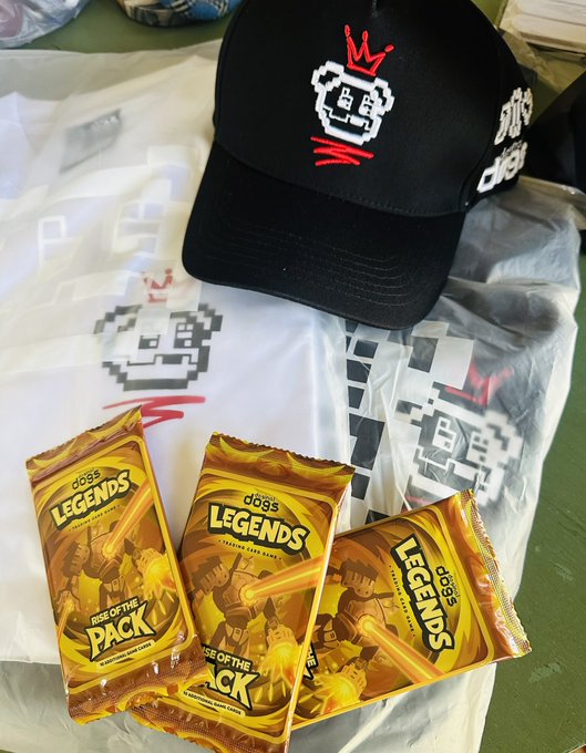 A black cap and a white t-shirt, both featuring a pixel-art character with a red crown logo, are displayed alongside three gold-colored packets labeled 'dogs LEGENDS RISE OF THE PACK'.