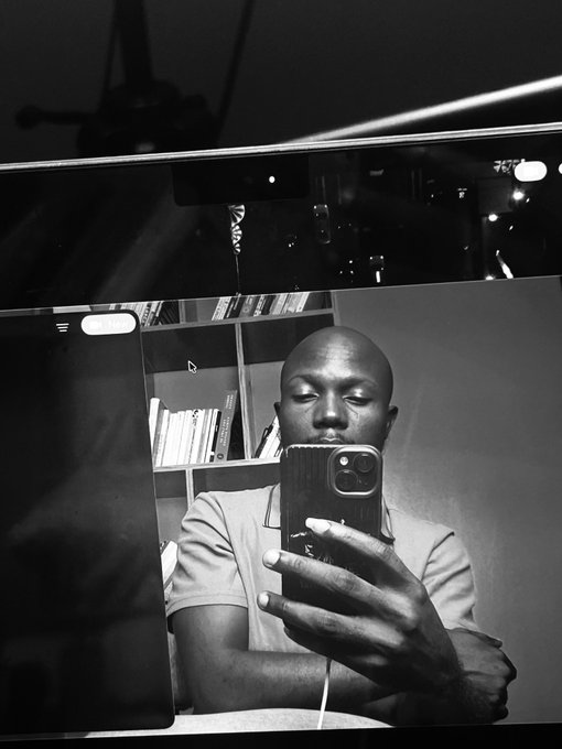 A black and white mirror selfie shows a young man holding up a phone to take the photo, with bookshelves and a computer monitor visible in the reflection behind him.