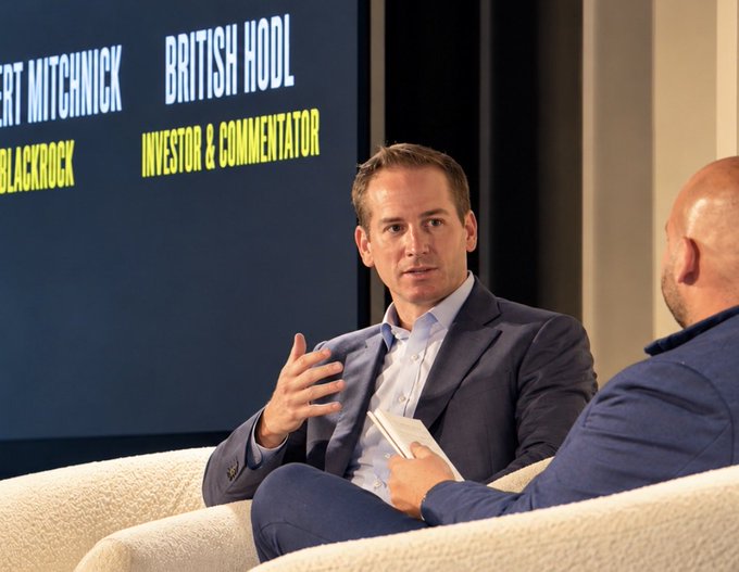 Two men in suits sit on a cream-colored couch during a panel discussion, with a screen behind them displaying text identifying "BRITISH HOOL" as an "INVESTOR & COMMENTATOR" alongside the name "MITCHNICK" and "BLACKROCK" — likely Robert Mitchnick, BlackRock's digital assets head, debating Bitcoin's market dynamics and future catalysts as referenced in the caption.
