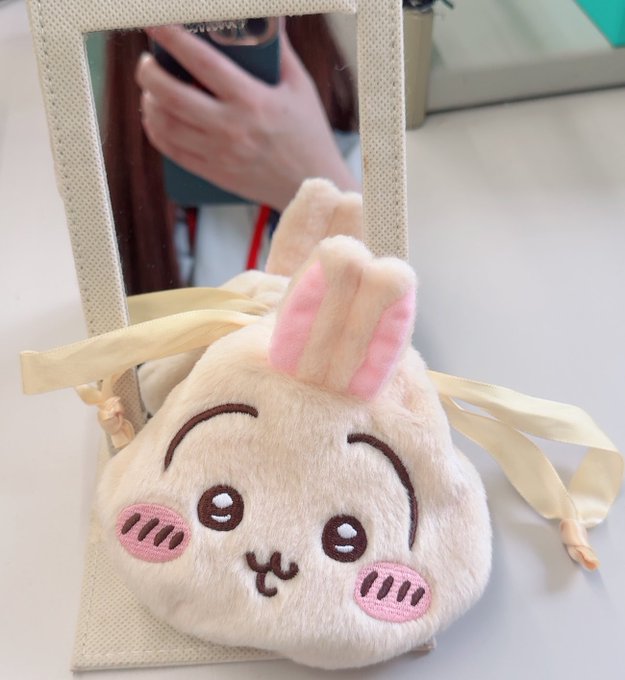 A plush rabbit handbag with a cute face design, tied with a ribbon, sitting in front of a mirror reflecting someone taking a photo with a phone.