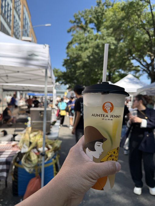 A hand holds an AUNTEA JENNY bubble tea cup at an outdoor market with white tents, trees, and pedestrians in the background.