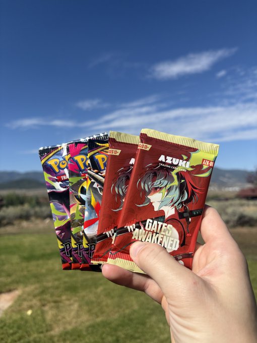 A hand holds up four colorful anime-themed collectible trading card packs against a bright blue sky with wispy clouds, including an 'AZUKI' branded pack labeled 'THE GATE: AWAKENED' and 'PoP' packs.