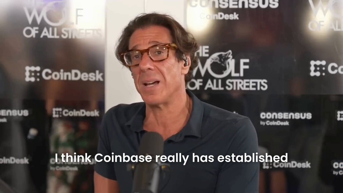 The image features a man wearing glasses standing at a microphone during an interview or discussion. He appears confident while addressing someone with a headset. The background of the scene has various text elements that suggest this could be related to Bitcoin or cryptocurrency news, such as "coinbase says Coinbase could be a generational company like Amazon or Microsoft" and "Focus on what is in the image and how it relates to the post".