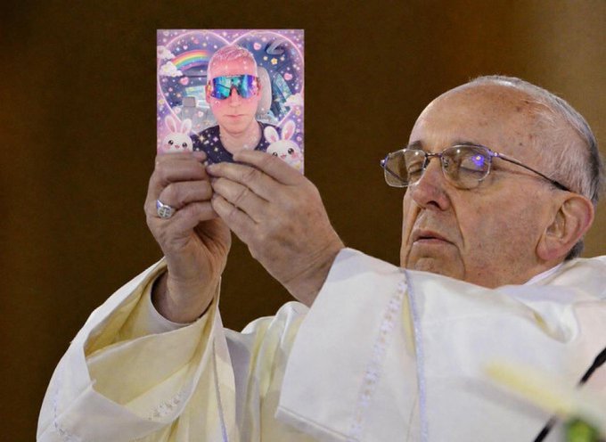 Pope Francis, wearing white robes and glasses, holds up a small card or photograph of a person wearing sunglasses, set against a galaxy-themed background with rainbows and bunnies.