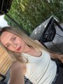 A selfie of a blonde woman in a white tank top outdoors with greenery in the background.