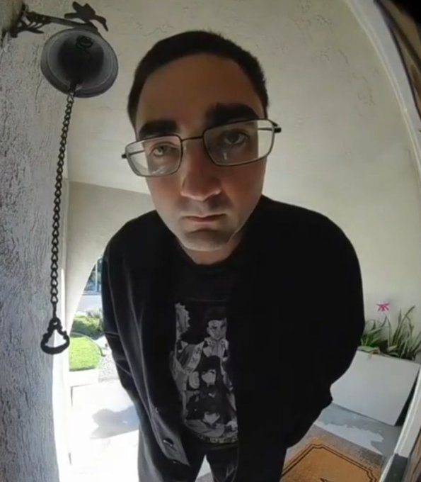 The image shows a man standing inside his home, wearing glasses, a black shirt with a picture of two cartoon characters printed on it, and looking down into a doorway or entryway. The entrance appears to be located at the bottom right corner of the photo. In this particular scene, there are three Ethereum holders visible in front of the man's eye level, implying that he might have recently purchased new Ether coins from them while entering his home. However, beyond those three holders, there is no further information about how these ETH wallets connect to the trading platform.