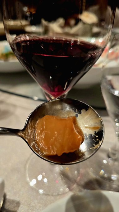 A close-up of a spoon holding an orange-colored paste held in front of a glass of red wine, suggesting a tasting or pairing experience.