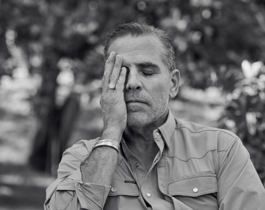 A black and white photograph of a man with gray hair and stubble wearing a button-up shirt, covering one eye with his hand in an outdoor setting.