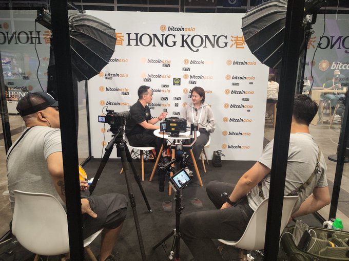 A live podcast or interview recording is underway at a Bitcoin Asia event in Hong Kong, with a man in a black shirt speaking into a microphone across from a woman, while camera equipment and two seated observers in the foreground frame the scene against the branded "Bitcoin Asia" and "HONG KONG" backdrop.