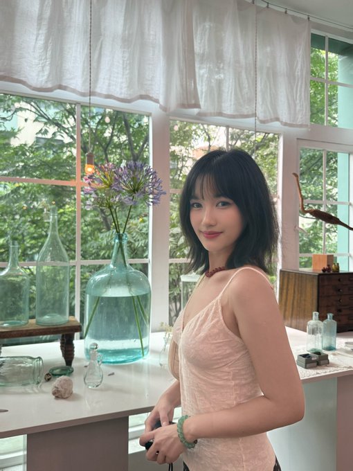 A young woman with short black hair wearing a light pink dress poses indoors near large windows, with glass bottles containing purple flowers on a table beside her.