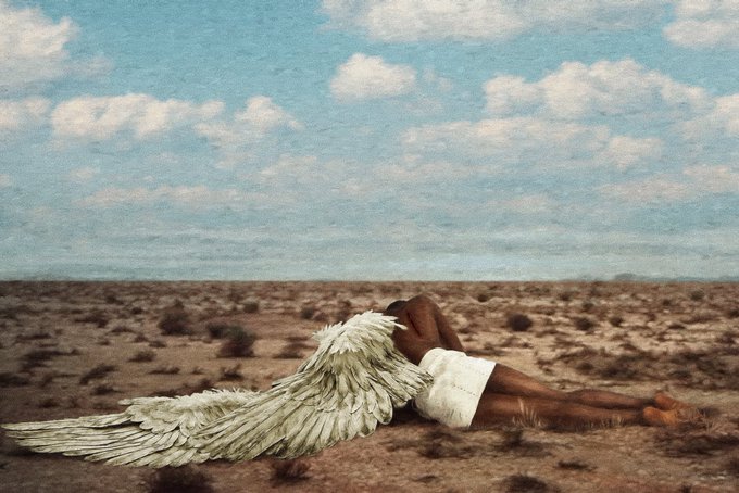 An artistic photograph of a Black figure with large white feathered angel wings, draped in white cloth, lying on a barren desert landscape under a blue sky with scattered clouds.