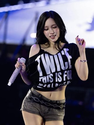 A young woman performs on stage holding a sparkly microphone, wearing a black crop top that reads 'TWICE THIS IS FOR' and denim shorts.