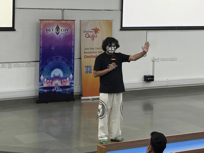 A presenter wearing a silver mask and casual clothing gestures with one hand raised while speaking at the Devcon SEA event, with banners for 'Devcon' and 'Onchain Guyju' visible behind them.