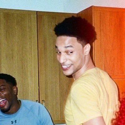 A young man with curly hair smiles at the camera while another person laughs in the foreground, both appearing to share a lighthearted moment. The casual indoor setting and lively expressions reflect the fun, social vibe associated with the "Aintsem" meme coin community featured in the post.