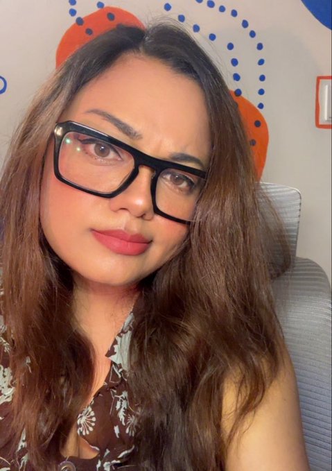 A woman with long brown hair and black-framed glasses takes a selfie in front of a colorful dotted backdrop, presumably promoting the Quip Network quests and statistics mentioned in her post about quantum-resistant crypto.