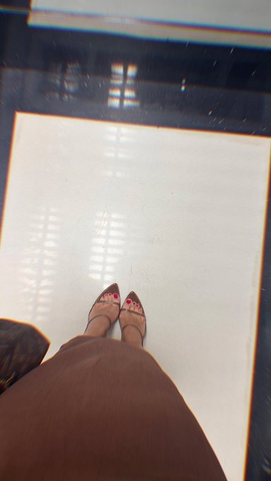 A first-person shot looking down at feet in strappy heels with red painted toenails, standing on a white surface and wearing a brown skirt—capturing a poised, "ready" moment before stepping out.