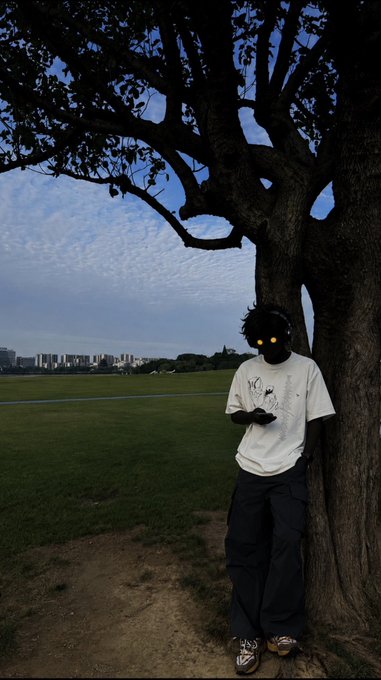 A young person wearing a graphic t-shirt and dark cargo pants leans against a large tree in an open field, their eyes glowing yellow in the dim twilight, with a distant city skyline on the horizon. The post "g m dawgs lets go again today" suggests a casual morning greeting to friends, paired with the moody, atmospheric photo to set a chill, ready-for-the-day vibe.