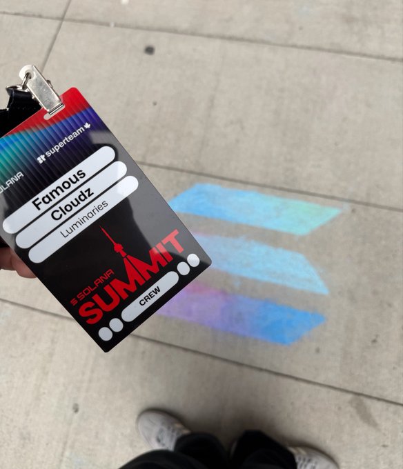 A hand holds a 'Solana Summit' badge for 'Famous Cloudz Luminaries Crew' with a holographic SuperteamUK lanyard, against a concrete floor with projected blue and purple light patterns.