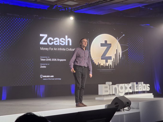 A man gives a presentation on a stage with a large screen displaying "Zcash Money For An Infinite Civiliz[ation]" and the Zcash logo, sponsored by BingX Labs and Zooko.