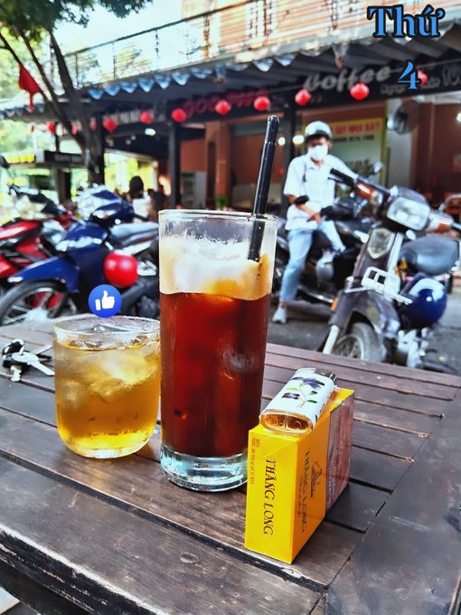Two iced drinks sit on a wooden table outside a café with red lanterns, alongside a yellow 'THANG LONG' cigarette pack, with motorbikes visible in the background.