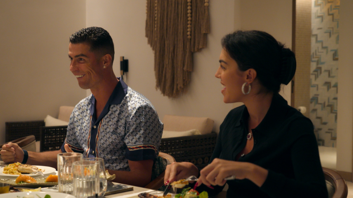 Cristiano Ronaldo sits at a dining table smiling alongside a woman in black attire, with food and wine glasses visible in the warmly lit restaurant setting.