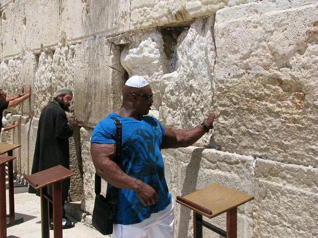 A man wearing a white kippah and blue shirt stands at the Western Wall with his hand pressed against the ancient stones, while another man in traditional Orthodox Jewish attire prays in the background. The image is juxtaposed with antisemitic text that mockingly references Jewish religious practices and pilgrimage, contradicting the solemn reality of the scene depicting visitors engaging in genuine prayer and reflection at one of Judaism's holiest sites.