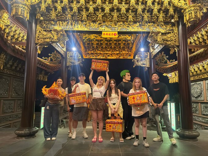 A group of seven young people pose in an ornate, traditional Chinese temple with golden decorations, holding decorative boxes with Chinese text including "永祥珠寶 30".