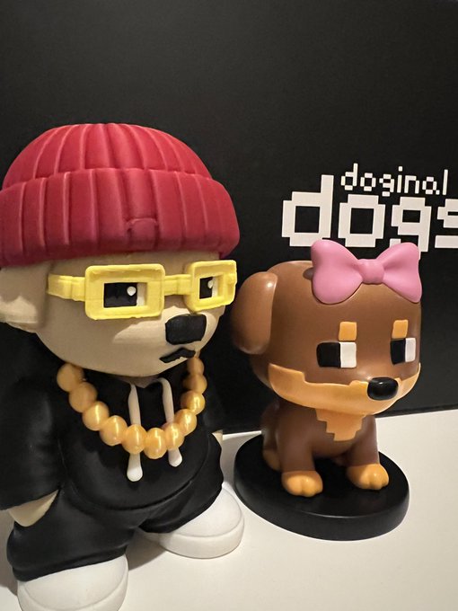 Two collectible figurines stand on a white surface against a dark background; one is a character wearing a red beanie, yellow pixelated glasses, and a gold chain, while the other is a brown dog-like figure with a pink bow, with 'doginal dogs' text visible behind them.