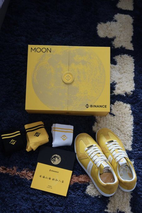 A Binance 'MOON' promotional box is displayed with matching yellow and white sneakers, three pairs of socks, and a card on a blue and white patterned rug.