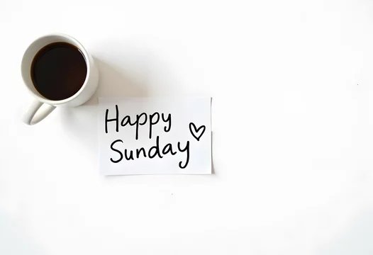 A flat lay of a white coffee cup beside a small note reading 'Happy Sunday ♡' on a white surface.
