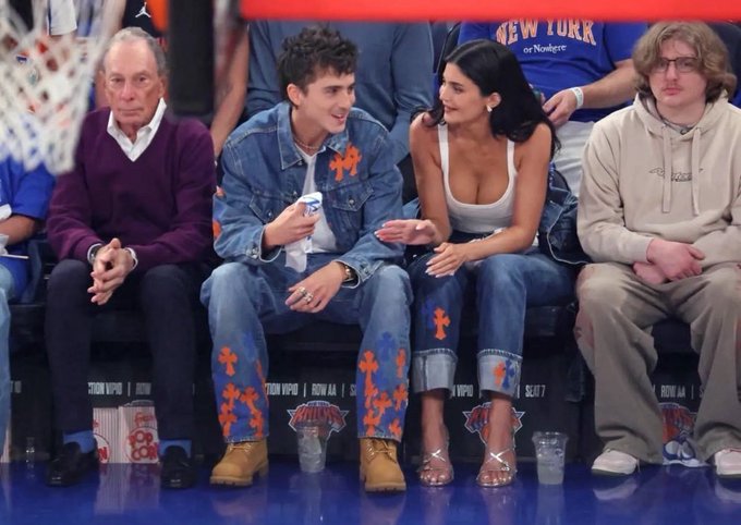 Four people sit courtside at a New York Knicks game; a man in a denim outfit with orange floral designs converses with a woman in a white top and jeans.