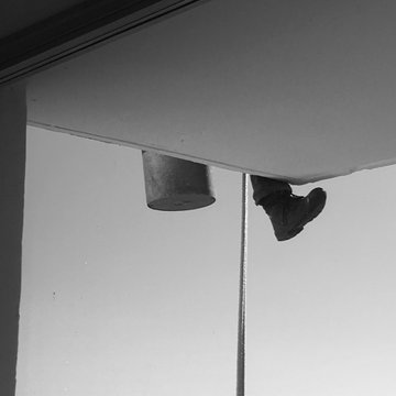 A black and white photograph showing a pair of feet in dark shoes dangling upside-down from the underside of a white overhang or platform against a plain wall.