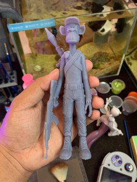 A person holds up a 3D-printed, unpainted figurine of a monkey character wearing a purple cap and carrying a rifle, with various painting supplies in the background.