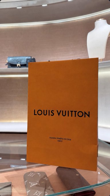 A vibrant orange Louis Vuitton shopping bag, prominently displaying "LOUIS VUITTON" and "MAISON FONDÉE EN 1854 PARIS," sits on a glass display in a luxury boutique, with additional monogram-patterned leather goods visible below—capturing a high-end shopping splurge funded by crypto gains.