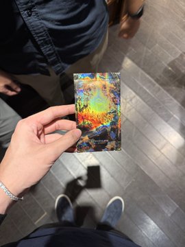 A hand holds up a vibrant, holographic collectible card against a tiled floor background, showcasing the colorful artwork as the person displays the small gift received at a crypto networking event. The card appears to be a promotional item from one of the project teams met during the BNB Chain gathering.