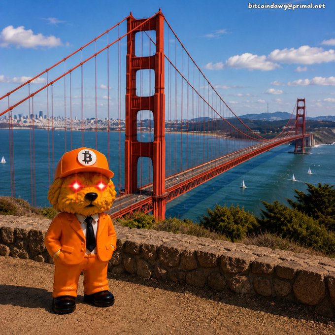 A teddy bear dressed in an orange suit and Bitcoin cap with glowing red eyes stands on a stone wall with the Golden Gate Bridge in the background, credited to bitcoindawg@primal.net.