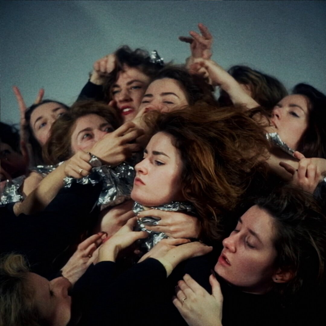 The image shows a group of people gathered together, with one woman wearing a scarf around her neck. The other women are holding onto her, creating an interesting scene that captures their attention. The photo appears to be taken during the day under natural light, as there is no visible artificial lighting in the picture. The caption at the bottom of the image reads "gm & happy tezostuesday, fam! my piece on @objktcom n o m a d e m e ( sound on ): 20".