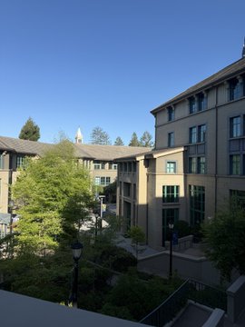 A view of a multi-story building with beige walls and large windows, surrounded by green trees under a clear blue sky, with a tower visible in the distance.