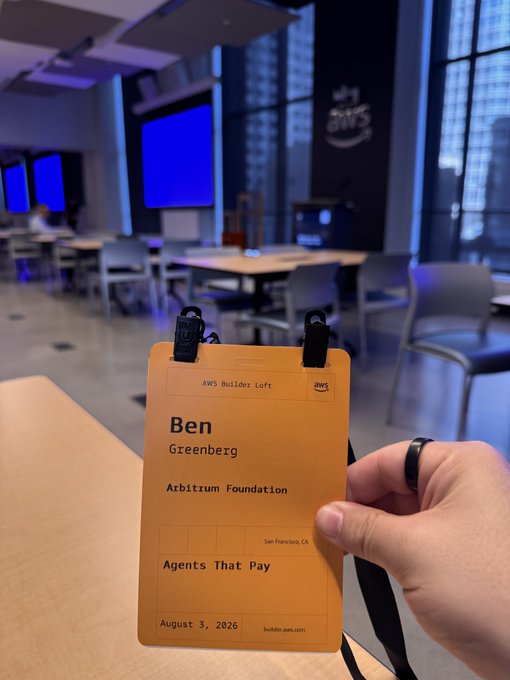 A hand holds an AWS Builder Loft attendee badge for 'Ben Greenberg' from 'Arbitrum Foundation', with the session 'Agents That Pay' dated August 3, 2026.