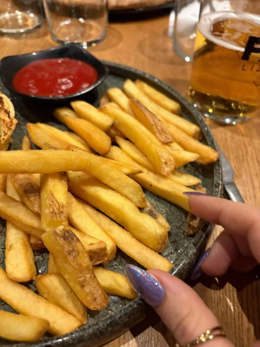 A plate of golden french fries served with a side of ketchup, with a hand featuring purple glittery nails pointing at the food, accompanied by a glass of beer.