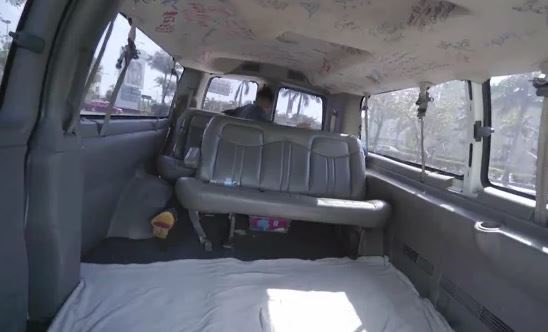 A complimentary shuttle van interior features a bed-like arrangement in the cargo area, decorated with handwritten messages and signatures across the ceiling and walls, while a passenger sits in the front seat as the vehicle transports them to Marina Bay Sands for the Token2049 conference in Singapore.